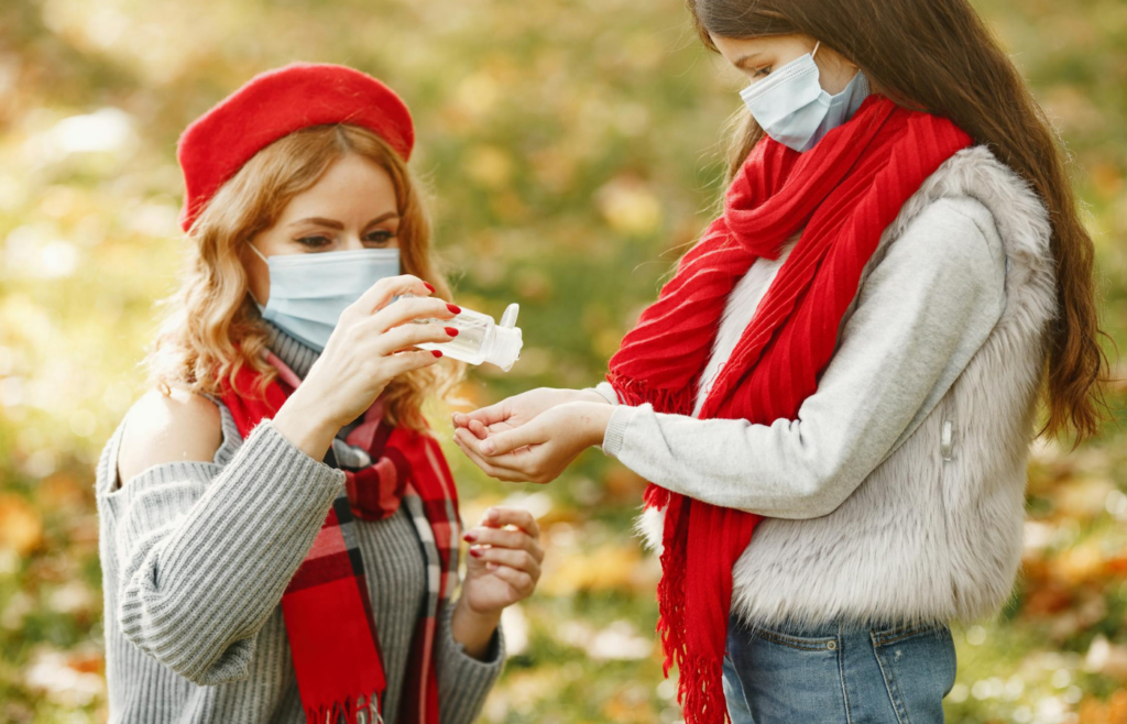 Two masked women using hand sanitizer in autumn park